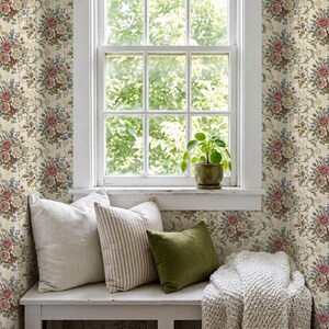 May include: A white wooden bench features three decorative pillows: one solid off-white, one beige and white striped, and one solid olive green. A small green potted plant sits on the windowsill against floral wallpaper with pink roses.