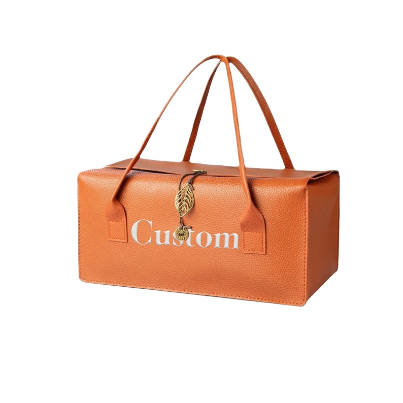 May include: A rectangular orange bag with a textured surface and two thin handles. The word Custom is printed in white on the front. A gold-colored metal leaf charm hangs from a thin cord at the center of the top edge of the bag.