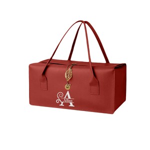 May include: A rectangular red textured bag with two thin handles. The front features a white monogram with a large letter A and the name Allan. A gold-colored metal leaf charm and a small circular pendant hang from the center closure.