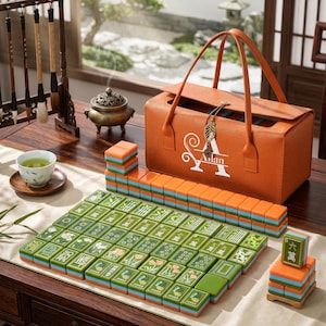 May include: A mahjong set featuring green tiles with floral and character designs, stacked in rows and columns. An orange leather-textured carrying case with the white text "Ailan" and a leaf-shaped charm sits behind the tiles on a wooden table.