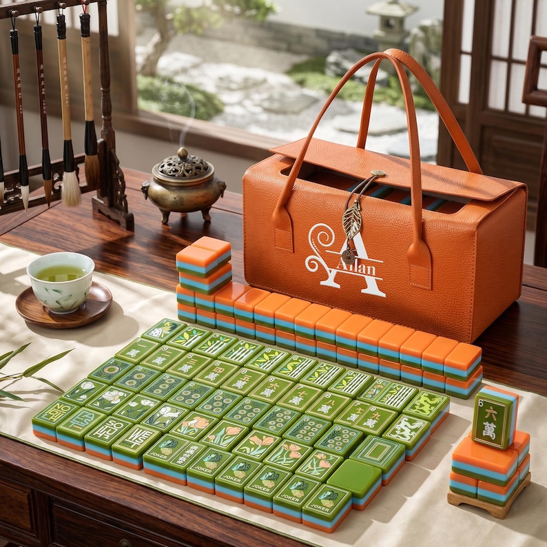 May include: A mahjong set featuring green tiles with floral and character designs, stacked in rows and columns. An orange leather-textured carrying case with the white text "Ailan" and a leaf-shaped charm sits behind the tiles on a wooden table.