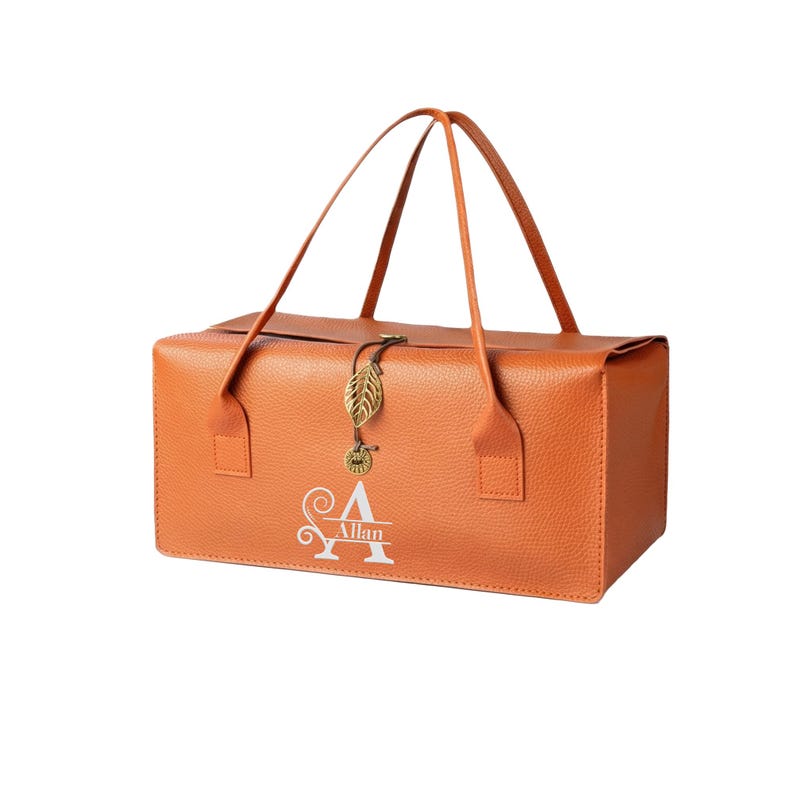 May include: A rectangular orange textured bag with two thin handles. The front features a white monogram with the name Allan. A gold-toned metal charm shaped like a leaf and a small circular pendant hang from the center closure of the bag.