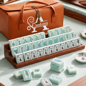 Puede incluir: Un juego de mahjong con fichas blancas que presentan ilustraciones verdes y negras, n&uacute;meros y caracteres chinos, dispuestas en un soporte de madera. Detr&aacute;s de las fichas hay un estuche de transporte naranja con una letra A blanca y la palabra Custom.