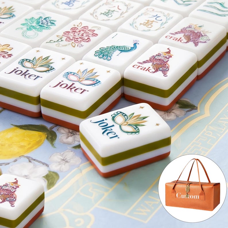May include: A set of rectangular mahjong tiles with white faces featuring colorful illustrations of masks, peacocks, flowers, and Chinese characters, including the word joker. Each tile has a layered green and orange base. A tan carrying bag is shown.