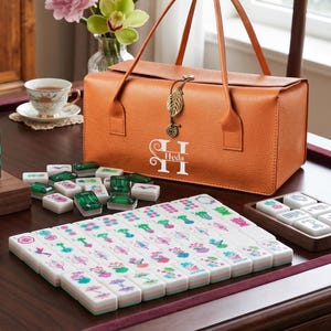 May include: An orange textured carrying case with a leaf charm and the text Heda, containing a mahjong set. The set includes white tiles with colorful floral and botanical illustrations, along with several translucent green tiles arranged on a table.