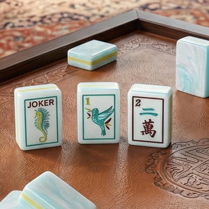 Puede incluir: Un conjunto de fichas de mahjong rectangulares con base blanca y bordes jaspeados en azul claro. Tres fichas est&aacute;n de pie, mostrando un caballito de mar con el texto JOKER, un colibr&iacute; con el n&uacute;mero 1 y el n&uacute;mero 2 con caracteres chinos.