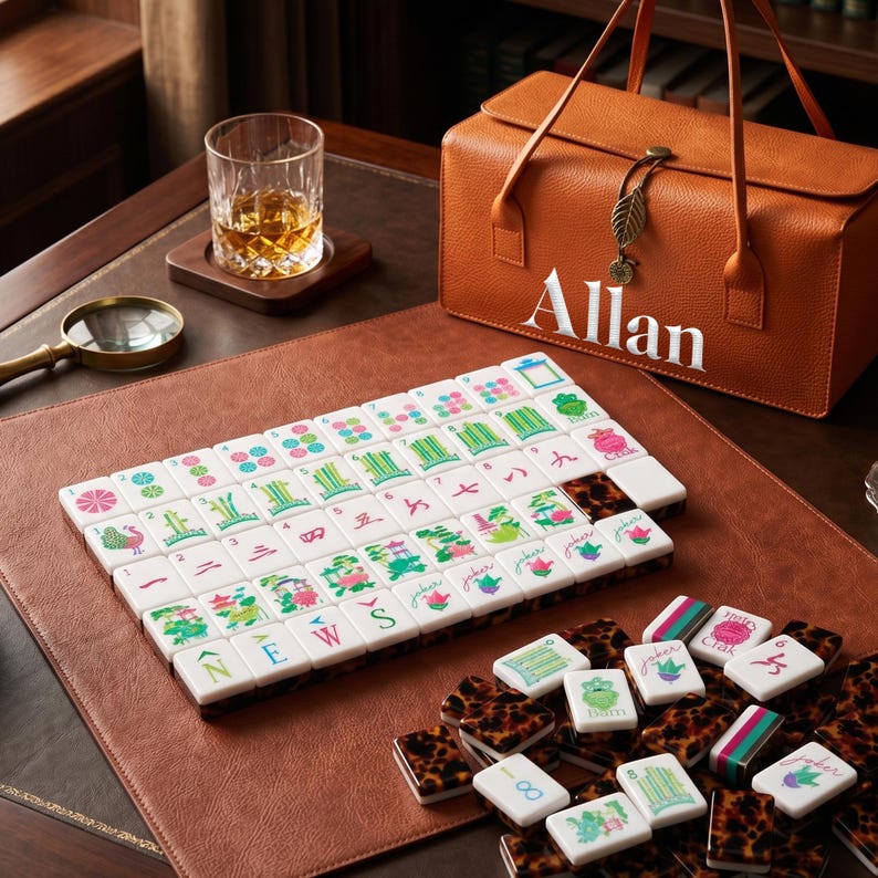 May include: A mahjong set featuring white tiles with colorful engraved symbols, numbers, and letters, alongside a tan leather carrying case with the name Allan embroidered in white, a glass of amber liquid, and a gold-rimmed magnifying glass.