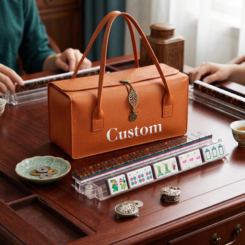 May include: A rectangular, orange, textured leather-like carrying case with two handles and a metal leaf charm, featuring the word "Custom" in white text. The case sits on a wooden surface alongside a clear plastic rack holding mahjong tiles.