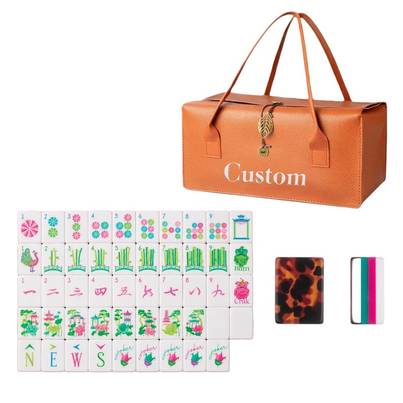May include: An orange rectangular carrying case with the word Custom printed in white, featuring a gold leaf charm. A set of white mahjong tiles with colorful floral, bamboo, and character designs, plus two additional tiles with tortoise and striped patterns.