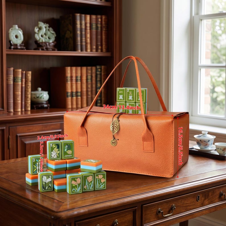 May include: An orange textured leather carrying case with two handles and a gold leaf charm, accompanied by a set of green and orange mahjong tiles. The case measures 34cm by 16.5cm, and individual tiles measure 2.4cm by 3.2cm.