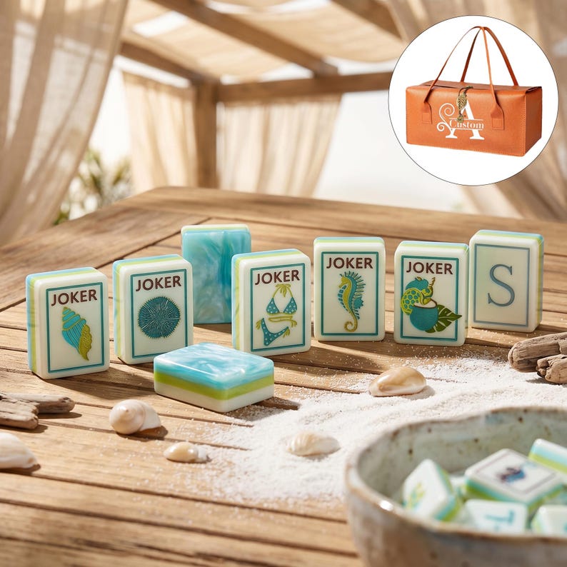 Puede incluir: Un juego de fichas de mahjong rectangulares con patrones jaspeados en blanco, azul y verde, ilustradas con conchas, un bikini, un caballito de mar y una tortuga, junto a una bolsa de transporte marr&oacute;n con el texto Custom y la letra A.