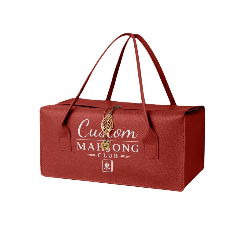 May include: A rectangular red textured bag with two thin handles. The front features white text reading Custom MAHJONG CLUB above a small square icon with the character 東. A gold-colored metal leaf charm hangs from the top center closure.