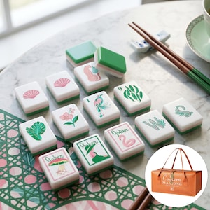 May include: A set of rectangular Mahjong tiles with white faces and green backs, featuring colorful illustrations of shells, flowers, birds, leaves, and the word Joker. A brown carrying bag with the text Custom Mahjong Club is also included.