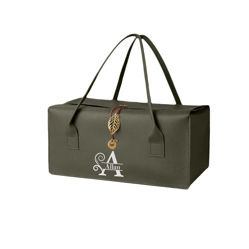 May include: A rectangular olive green bag with a textured surface, two thin handles, and a gold-toned leaf charm attached to a brown cord. The front features a white monogram design with a large letter A and the name Allan printed across it.
