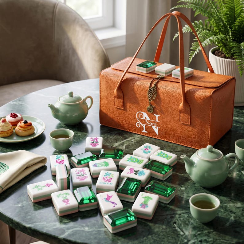 May include: A rectangular orange textured carrying case with a leaf-shaped charm and the name Nora printed in white, accompanied by a set of mahjong tiles featuring green translucent backs and various floral or bird designs, plus two light green teapots.