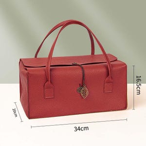 May include: A rectangular red handbag with a textured surface, two top handles, and a flap closure secured by a cord with a leaf and circular charm. The bag measures 34 cm in length, 20 cm in width, and 16.5 cm in height as indicated by labels.