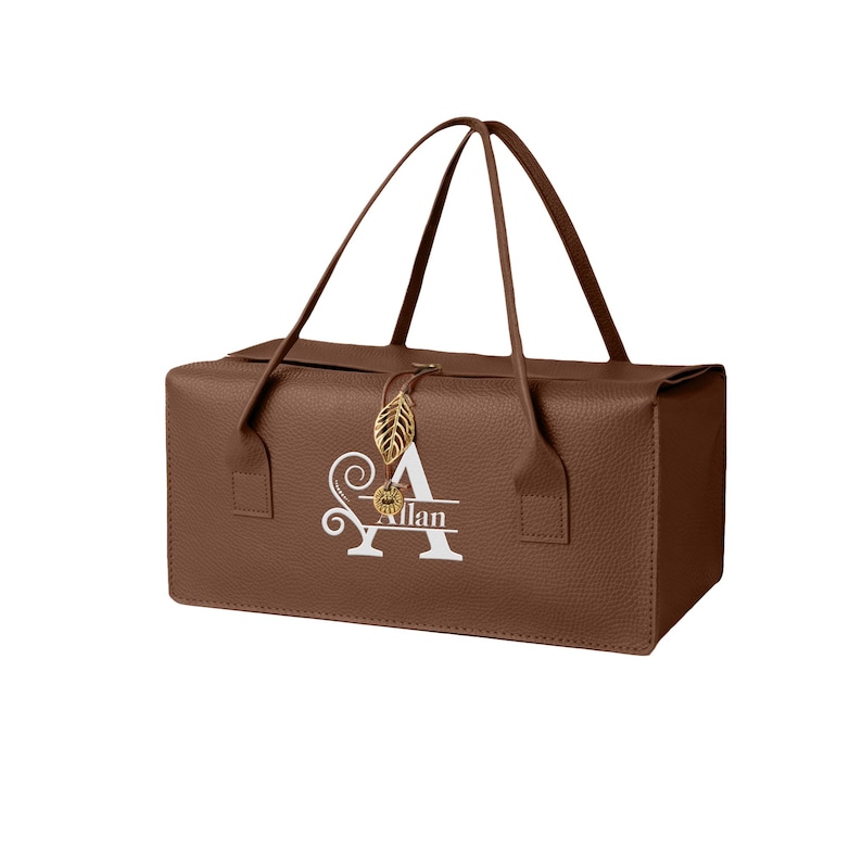 Puede incluir: Un bolso rectangular marr&oacute;n con superficie texturizada, dos asas finas y un dije dorado en forma de hoja. La parte frontal muestra una letra A grande en blanco con el nombre Allan impreso en el centro con una tipograf&iacute;a cursiva blanca.