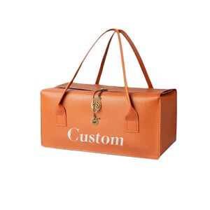 May include: A rectangular orange bag with a textured surface and two thin handles. The word Custom is printed in white on the front. A gold-colored metal leaf charm and a small circular pendant hang from a cord attached to the center top edge.