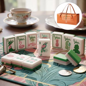 May include: A set of white mahjong tiles featuring green and pink illustrations of jokers, birds, and tropical motifs, accompanied by a rectangular orange carrying case with the text Custom Mahjong Club printed on the front surface.