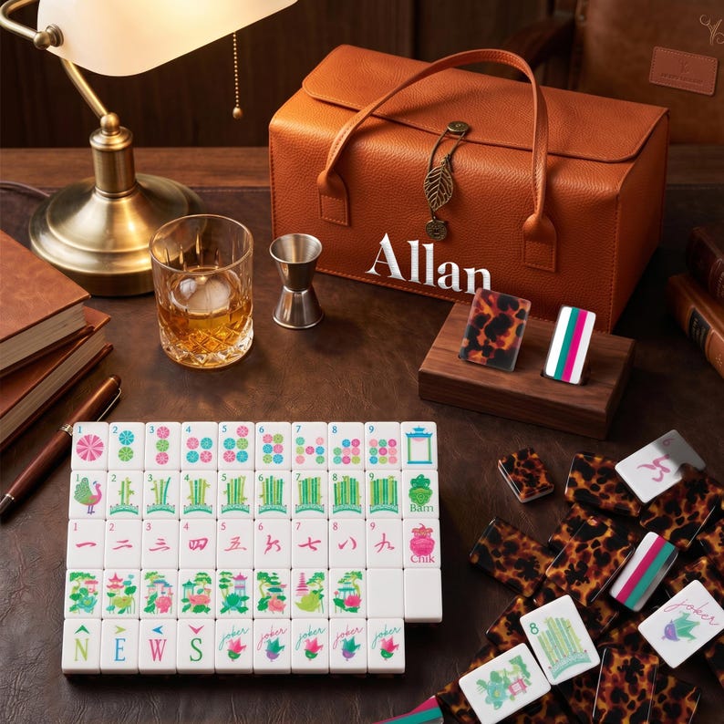 May include: A brown leatherette carrying case with the name Allan printed in white, accompanied by a set of mahjong tiles featuring colorful illustrations, numbers, and Chinese characters, alongside a glass tumbler and a metal jigger.