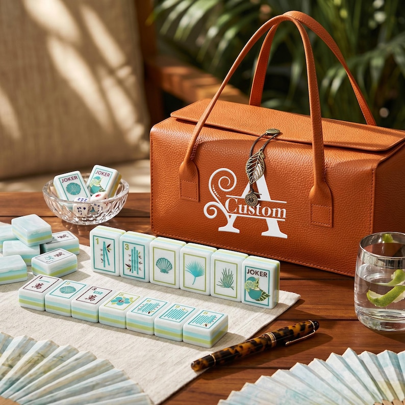 Puede incluir: Un malet&iacute;n de transporte rectangular de color naranja con textura, asa y un dije en forma de hoja, con una letra A blanca y la palabra Custom. Fichas de mahjong con ilustraciones verdes y azules dispuestas sobre una tela, junto a un vaso de agua y un bol&iacute;grafo.