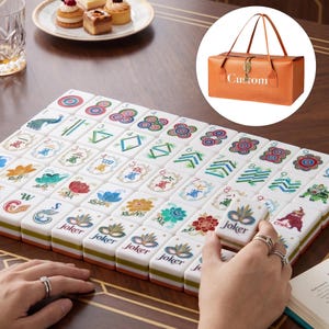May include: A set of white mahjong tiles with colorful floral and numerical designs, including tiles labeled Joker with a mask graphic. An orange rectangular carrying case with the text Custom is shown in a circular inset above the tiles.
