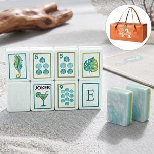 Puede incluir: Un juego de fichas de mahjong rectangulares con caras blancas que presentan ilustraciones en verde y azul de un caballito de mar, conchas, una hoja de monstera, la letra E y una copa de c&oacute;ctel con la etiqueta JOKER. Dos fichas muestran un patr&oacute;n marmoleado.