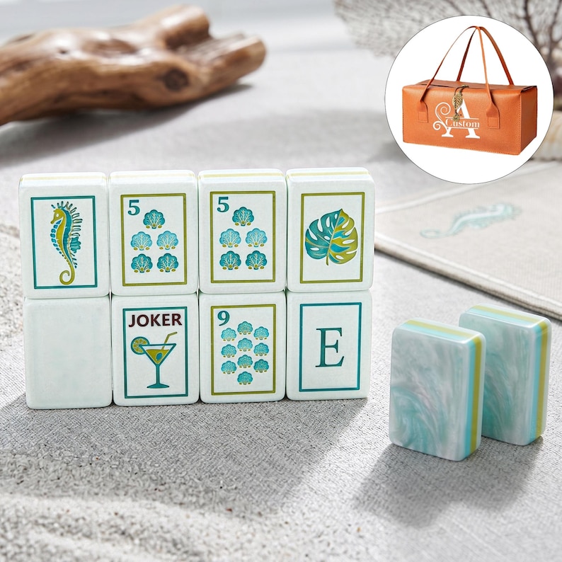 Puede incluir: Un juego de fichas de mahjong rectangulares con caras blancas que presentan ilustraciones en verde y azul de un caballito de mar, conchas, una hoja de monstera, la letra E y una copa de c&oacute;ctel con la etiqueta JOKER. Dos fichas muestran un patr&oacute;n marmoleado.