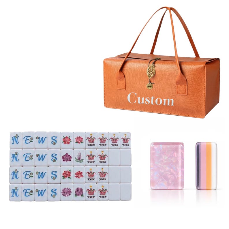 May include: An orange rectangular carrying case with the word Custom printed in white, featuring two handles and a gold leaf charm. Included are white mahjong tiles with blue letters, floral designs, and joker icons, plus two translucent tiles.