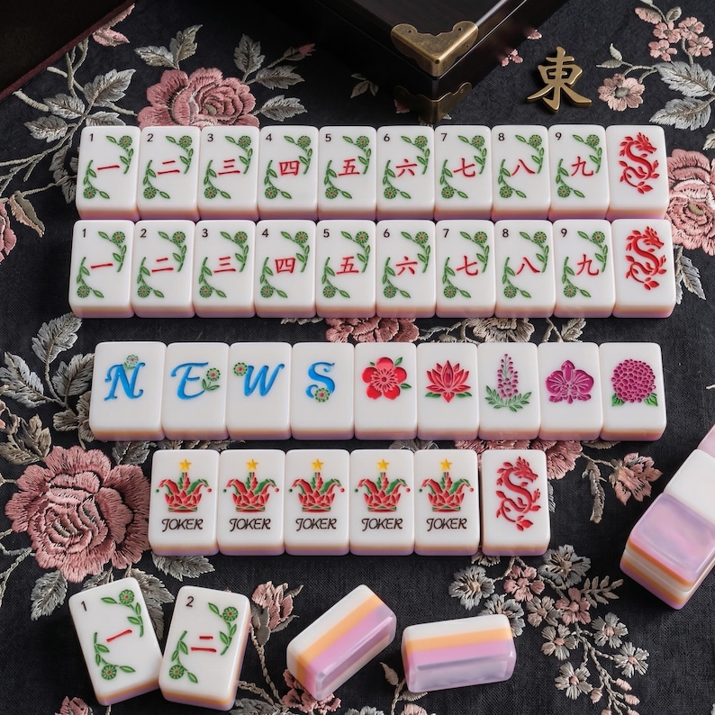 K&ouml;nnte beinhalten: Ein Set rechteckiger Mahjong-Spielsteine mit wei&szlig;er Oberfl&auml;che und abgerundeten Kanten. Die Steine zeigen rote chinesische Ziffern von eins bis neun, rote Drachensymbole, blaue Buchstaben f&uuml;r NEWS, bunte Blumenillustrationen und Joker-Steine mit Kronen.