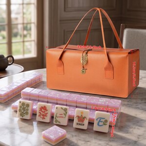May include: An orange rectangular carrying case with a gold leaf charm and two handles, measuring 13.4 inches long and 6.5 inches high. In front are several mahjong tiles with pastel pink and white layers, featuring various floral and character designs.