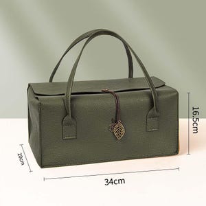May include: A rectangular olive green handbag with a textured surface, two top handles, and a flap closure secured by a brown cord with a metal leaf charm. Dimensions are 34 cm in length, 20 cm in width, and 16.5 cm in height as indicated in the image.