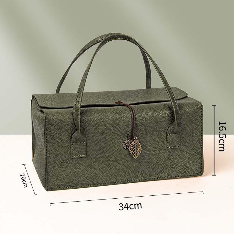 May include: A rectangular olive green handbag with a textured surface, two top handles, and a flap closure secured by a brown cord with a metal leaf charm. Dimensions are 34 cm in length, 20 cm in width, and 16.5 cm in height as indicated in the image.