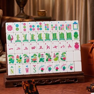 May include: A set of white Mahjong tiles arranged in five rows on a dark wooden display stand. The tiles feature colorful illustrations of flowers, bamboo, Chinese numerals, the letters N, E, W, S, and the word Joker with a floral graphic.