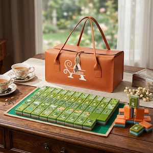 May include: A tan leather carrying case with a leaf-shaped charm and the letter A printed on the front, accompanied by a set of green and orange mahjong tiles arranged on a green mat, with a small stack of tiles placed to the right side.