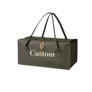 May include: A rectangular olive green bag with a textured surface and two thin handles. The word Custom is printed in white on the front. A brown cord with a gold-colored leaf-shaped charm and a small circular pendant hangs from the top center.