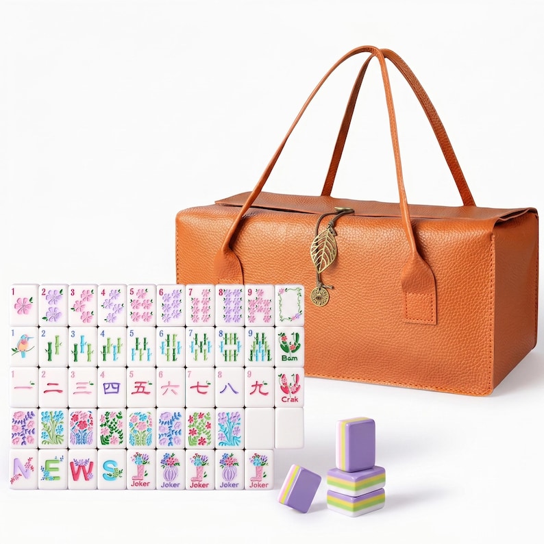 May include: A mahjong set featuring white tiles with colorful floral, bamboo, and character designs, including tiles labeled NEWS, Joker, and Crak. The set includes a textured orange carrying bag with a gold leaf charm and a stack of purple and green tiles.