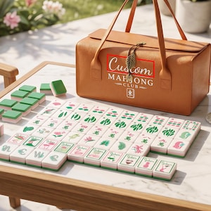May include: A tan leather carrying case with the text Custom MAH JONG CLUB and a leaf charm, accompanied by a set of rectangular game tiles featuring green, pink, and white designs, including various floral, joker, and numerical symbols.