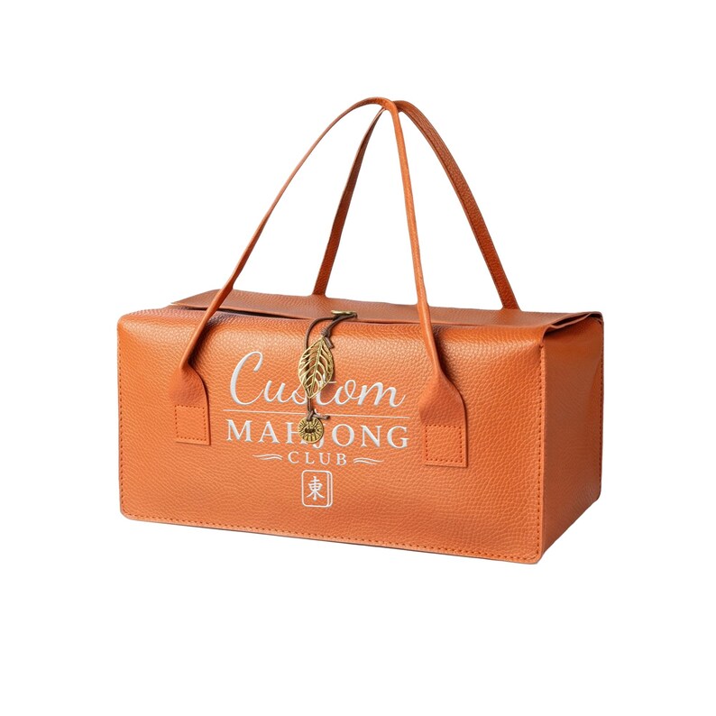 May include: A rectangular orange textured bag with two thin handles. The front features white text reading Custom MAHJONG CLUB and a small square icon with the character 東. A gold-toned metal leaf charm hangs from the center closure.