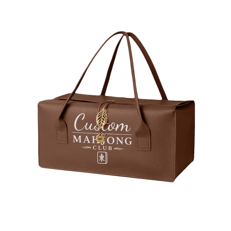 May include: A brown rectangular carrying case with two handles, featuring the white text Custom MAHJONG CLUB and a square symbol with a character. A gold-colored leaf-shaped charm hangs from the center of the top edge of the bag.