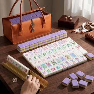 Op de afbeelding: Een mahjongset met witte tegels voorzien van kleurrijke bloem- en karakterontwerpen, naast effen lavendelkleurige tegels. De set bevat een goudkleurig metalen tegelrek, een gestructureerde oranje draagtas met een bladbedel en een donkere houten doos.