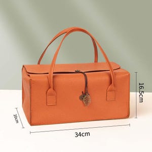May include: A rectangular orange handbag with a textured surface, two top handles, and a flap closure secured by a thin cord with a metal leaf charm. The bag measures 34 cm in length, 20 cm in width, and 16.5 cm in height as indicated by labels.