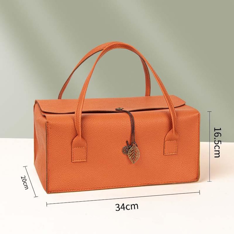 May include: A rectangular orange handbag with a textured surface, two top handles, and a flap closure secured by a thin cord with a metal leaf charm. The bag measures 34 cm in length, 20 cm in width, and 16.5 cm in height as indicated by labels.