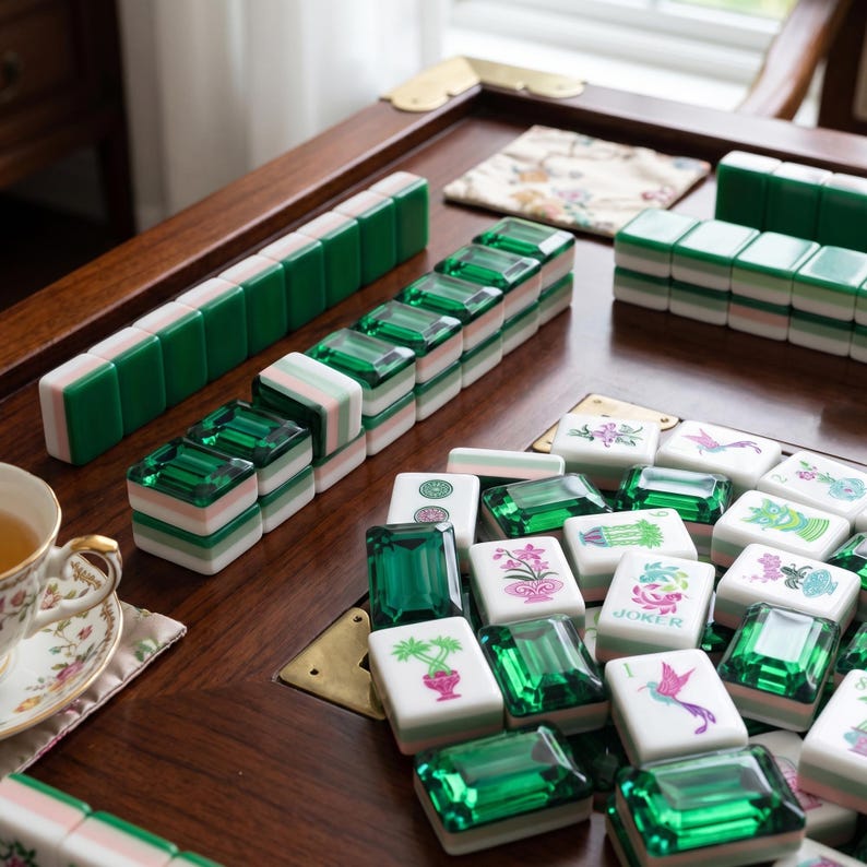 May include: A mahjong set featuring white tiles with green translucent backs and colorful floral or bird illustrations, arranged on a wooden table alongside a teacup and saucer with a floral pattern and a small decorative fabric square.