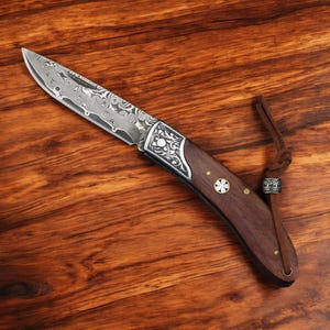 May include: A folding knife with a patterned steel blade and a dark wood handle. The handle has decorative metal accents and a leather lanyard. The knife is lying on a wooden surface.
