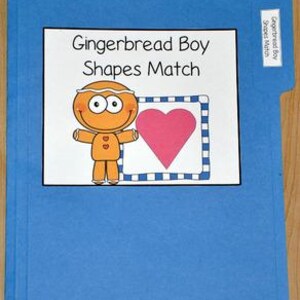 Puede incluir: Una carpeta azul con una etiqueta blanca que dice Gingerbread Boy Shapes Match. La etiqueta muestra un personaje de pan de jengibre junto a un gran corazón rosa dentro de un borde cuadrado a cuadros azules y blancos.