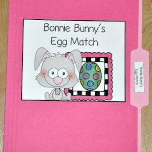 May include: A pink file folder with a white label featuring the text "Bonnie Bunny's Egg Match." The label includes a cartoon bunny and a decorated Easter egg. The folder is on a wooden surface.