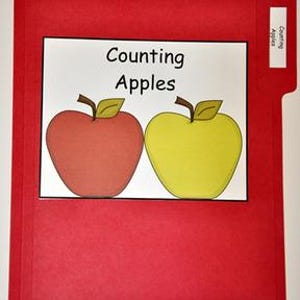 May include: A red file folder with a white rectangle containing the words "Counting Apples" above two apple illustrations. One apple is red, the other is yellow. A small tab on the right side reads "Counting Apples".