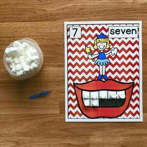 Può includere: Un contenitore di plastica trasparente pieno di mini marshmallow, una penna blu e una scheda didattica colorata. La scheda presenta il numero "7" e la parola "seven", insieme a un'illustrazione di un cartone animato di una fatina dei denti e una grande bocca con denti di marshmallow.
