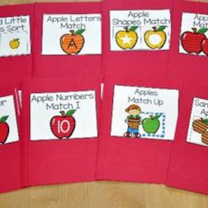 Può includere: Una collezione di cartelle rosse, ciascuna etichettata con attività educative a tema mela. I titoli includono "Big and Little Apples Sort", "Apple Letters Match" e "Apple Shapes Match". Le cartelle sono disposte su una superficie marrone chiaro.